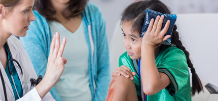 Child with dark hair and a green shirt holding head with their head from an injury. An adult in the background holding up 4 fingers pointed at the child to initiate concussion testing.